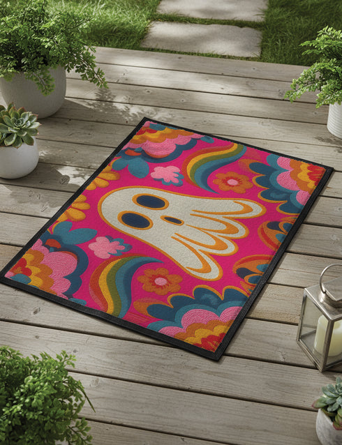 A rectangular polyester doormat with a vibrant, psychedelic design on a hot pink background. In the center is a cute, cartoonish ghost in off-white with yellow and orange outlines; it has large, dark, expressive eyes. The ghost is surrounded by a maximalist pattern of 70s-style floral motifs and puffy clouds in shades of teal, blue, orange, and light pink. Two small, colorful rainbows arc over the ghost's shoulders. The doormat has a durable, textured surface and is framed by a sleek black trim.